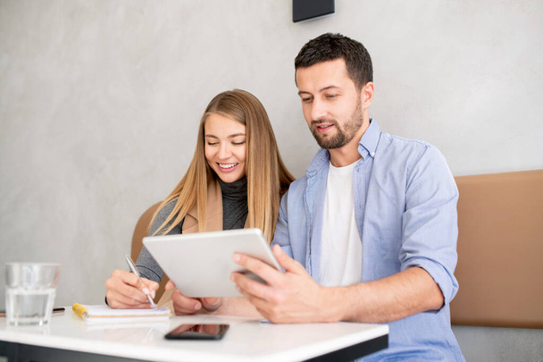 junger Geschäftsmann mit Touchpad blättert durch Online-Informationen, während sein hübscher Kollege bei einem Meeting Notizen macht - Foto, Bild