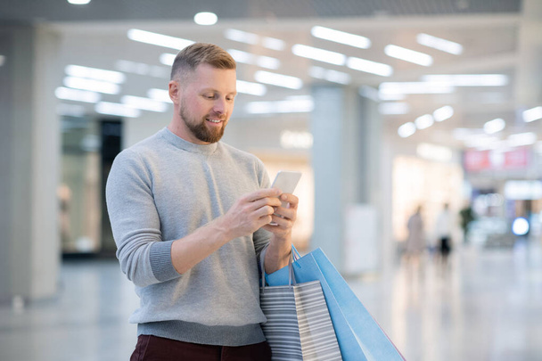 Молодой человек с бумажными пакетами и смартфоном смотрит на промо на экране или просматривает страницы интернет-магазина, стоя в торговом центре
 - Фото, изображение