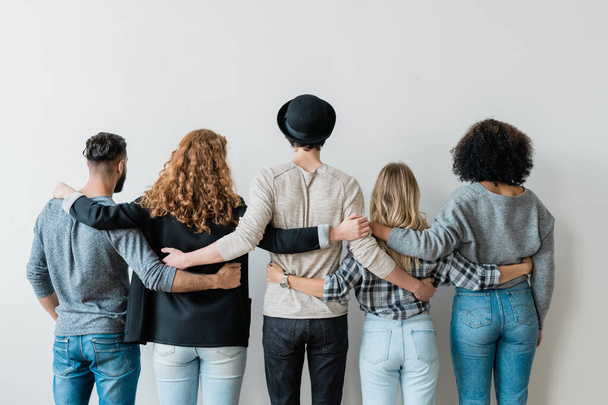 Rückansicht der Reihe junger lässiger College-Freunde, die sich umarmen, während sie vor einer weißen Wand stehen - Foto, Bild