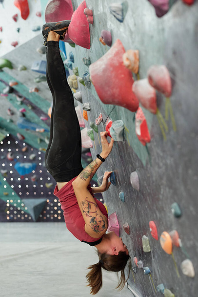 Jeune sportive qualifiée suspendue à l'envers avec ses pieds et ses mains tenant par des rochers artificiels sur un mur d'escalade
 - Photo, image