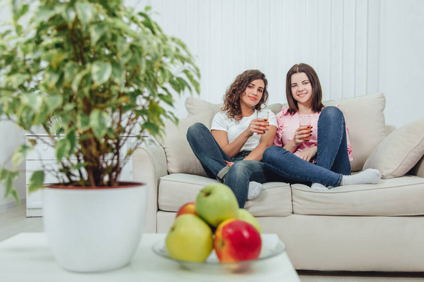 Blurred plate of apples and flower pot on the forefront. Two cute young girls sitting on the sofa with glasses of juice in hands, on the background - Photo, image