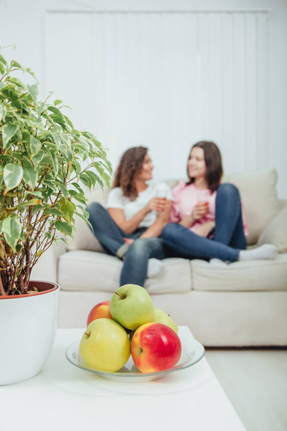 Plate of apples and flower pot on the forefront. Two cute young girls sitting on the sofa with glasses of juice in hands, communicating, on the blurred background - Foto, immagini