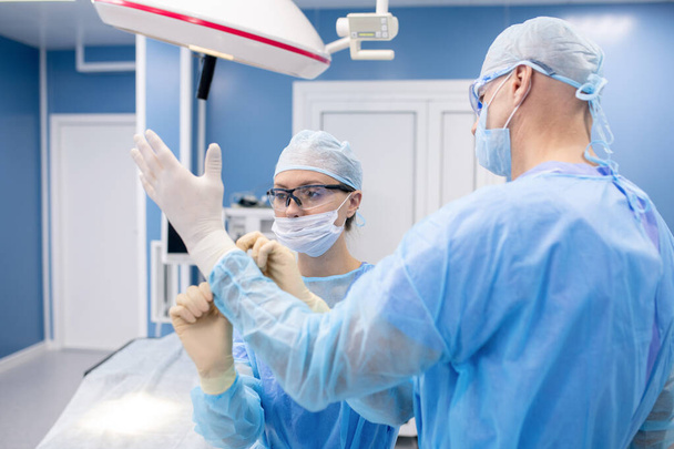 Νεαρή βοηθός με στολή που βοηθάει χειρουργό να φορέσει ιατρικά γάντια πριν χειρουργηθεί στο νοσοκομείο - Φωτογραφία, εικόνα