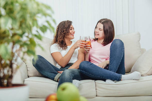 Blurred plate of apples and flower pot on the forefront. Two cute young girls sitting on the sofa clinking glasses with juice on the background. - Photo, image
