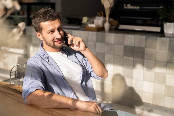 Szczęśliwy młody biznesmen ze smartfonem rozmawiając z kolegą lub konsultantem klienta podczas relaksu przy stole w kawiarni - Zdjęcie, obraz