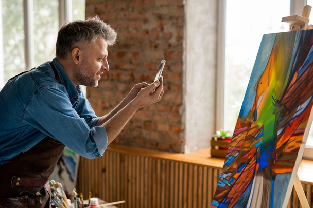 Seitenansicht des glücklichen Künstlers mittleren Alters mit Smartphone beim Fotografieren abstrakter Kunstwerke auf Staffelei in seinem Atelier - Foto, Bild