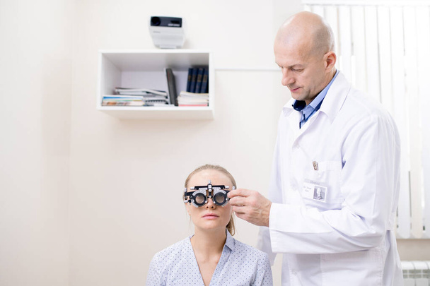 Bald professional ophthalmologist examining eyesight of young female patient to get her individual lens for eyewear - Foto, Imagen