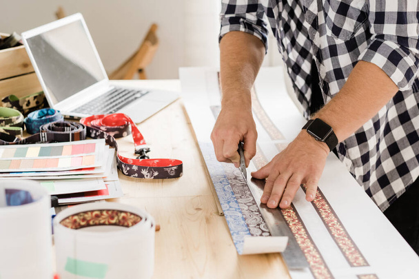 Young casual craftsman with knife and ruler bending over table while cutting decorative workpieces for creative pet collars - Foto, Bild