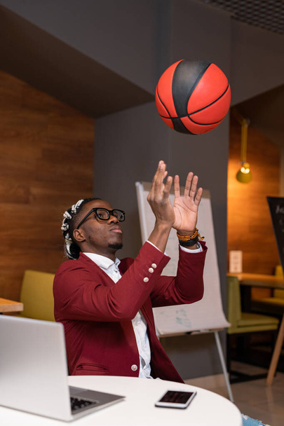 African guy in smart casualwear throwing ball upwards while sitting by table in modern cafe with laptop in front - Fotoğraf, Görsel