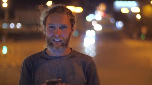 Close-up Portret van een knappe blanke bebaarde, langharige jongeman met smartphone die iemand uitnodigt door gebaren en gezichtsuitdrukkingen. Onscherpe avondverlichting van een stad op de achtergrond - Video