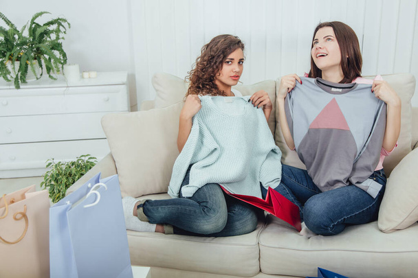 Young girls came back from the shop. They are sitting on the sofa, taking clothes out from the bags and trying it on. Expressive satisfied face expressions. - Photo, image