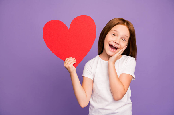 Photo of pretty overjoyed little foxy lady hold large red paper heart figure classmate boy made holiday present surprise wear white t-shirt isolated purple background - Foto, Imagem