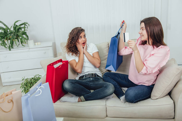 Friends sitting on the couch. One girl is smiling, holding purchase in blue shop bag and credit card, another is looking at her with surprised even confused face expression. - Foto, Imagem