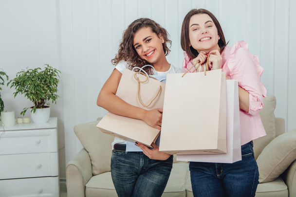 Happy girls are at home after shopping. They are smiling, standing shoulder to shoulder hugging shop bags. - Photo, image