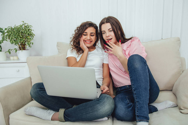 Excited girls sitting on the sofa with crossed legs watching film on the laptop. - Zdjęcie, obraz