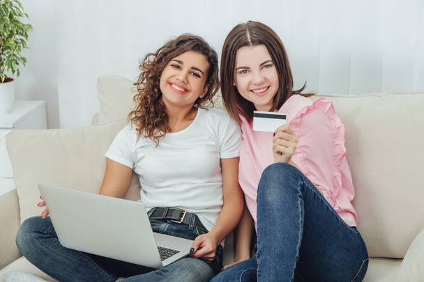 Kaksi onnellista naispuolista ystävää istuu sohvalla kannettavan tietokoneen ja luottokortin avulla ostoksia varten verkossa
. - Valokuva, kuva