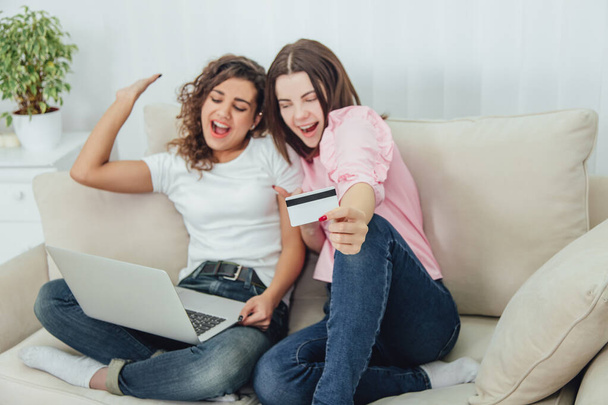 Extremely joyful shopaholic girls sitting on the sofa, with satisfied face expressions, using laptop and card for shopping online. - Foto, imagen
