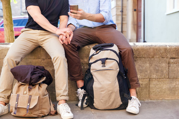 Close Up Of Male Gay Couple On Vacation Sitting On Wall With Backpacks Looking At Mobile Phone - Фото, зображення
