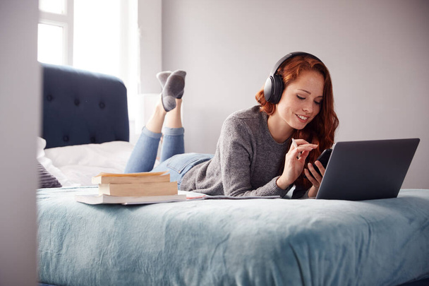 Kobieta studentka nosząca słuchawki leży na łóżku we wspólnym domu z telefonem komórkowym przy użyciu laptopa - Zdjęcie, obraz