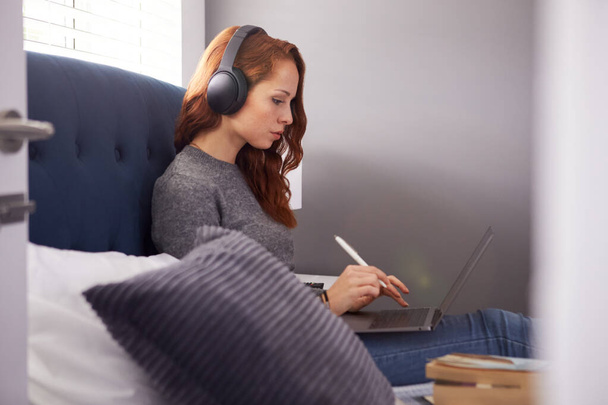 Студент коледжу носить бездротові навушники сидячи на ліжку у спільному будинку за допомогою ноутбука
 - Фото, зображення