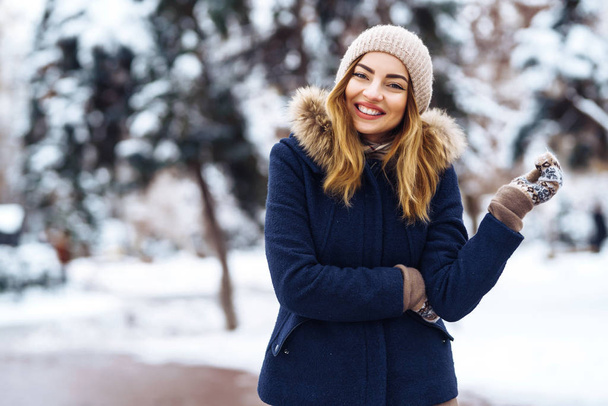 冬の森の雪の木の中に美しい女性が立っている。青いジャケットを着て帽子とミトンを編んだ若い女の子。幸せな冬の時間。冬の公園でファッションの若い女性。クリスマス.  - 写真・画像