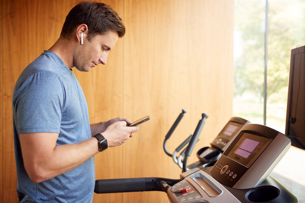 Man Exercising On Treadmill At Home Wearing Wireless Earphones Checking Mobile Phone - Valokuva, kuva