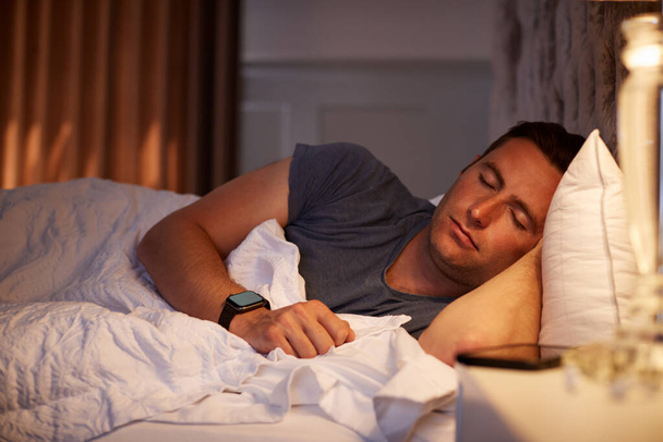 Man Sleeping In Bed Wearing Smart Watch Illuminated By Bedside Lamp - Photo, Image