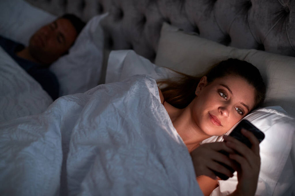 Woman Lying In Bed Checking Mobile Phone Whilst Man Sleeps Next To Her - Foto, immagini