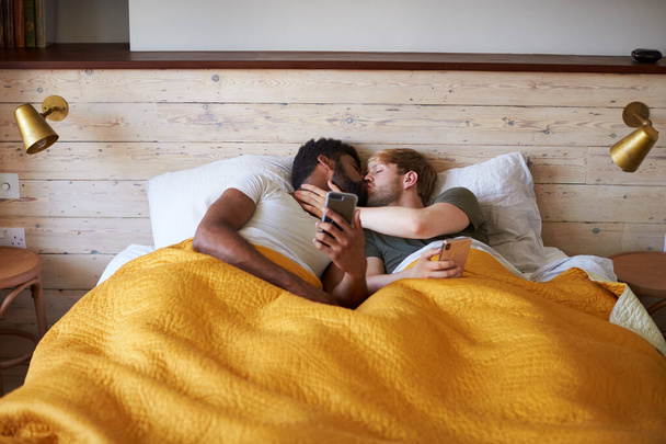 Male Gay Couple Lying In Bed At Home Checking Mobile Phones Toge - 写真・画像