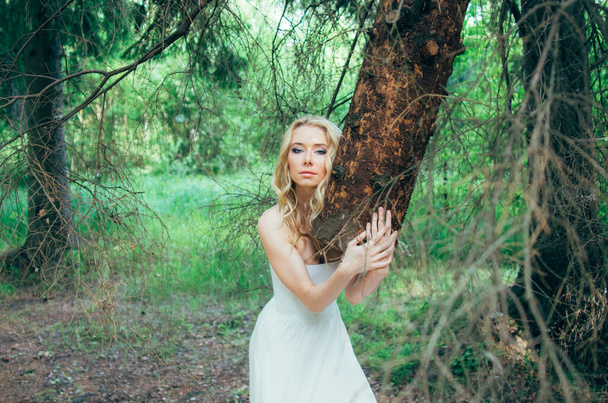 Portrait of young blond bride in forest holding tree trunk - 写真・画像