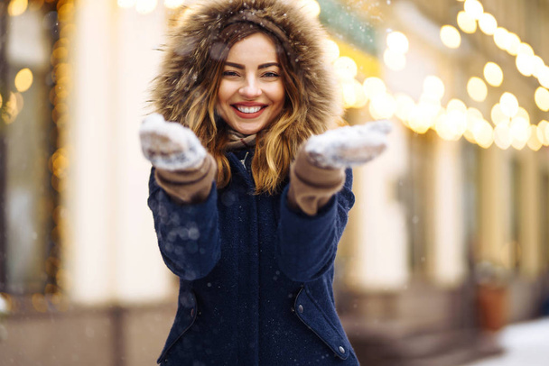 Beautiful  girl in a blue jacket and knitted hat and mittens posing in street of city. Garland lights. Enjoying snow, smiling to camera, cheerful  new year mood, true emotions. Winter fashion. - Фото, изображение