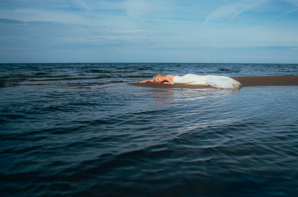 Portret van jonge blonde bruid ligt op zand tussen het water - Foto, afbeelding