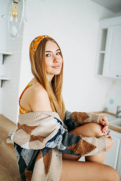 Belle sensuelle souriante jeune femme belle fille aux cheveux longs portant dans le confortable cardigan tricoté assis sur la table de cuisine à la maison, intérieur lumineux
 - Photo, image