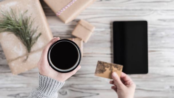 Woman with cup of coffee, credit card and tablet. Packing gifts. Cardboard box in craft paper on the rustic wood planks background. Christmas and New Year concept.  - Foto, Imagem