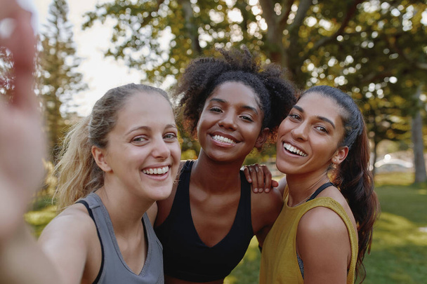 Smiling portrait of diverse female friends looking at camera taking selfie in park - fitness friends taking a selfie after exercising  - 写真・画像