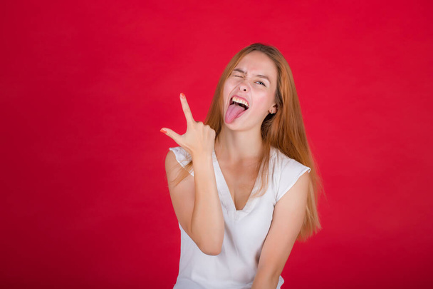 Portrait of a nice attractive cheerful red-haired girl in a white blouse shows tongue and two fingers on her right hand isolated on a bright red background - Φωτογραφία, εικόνα