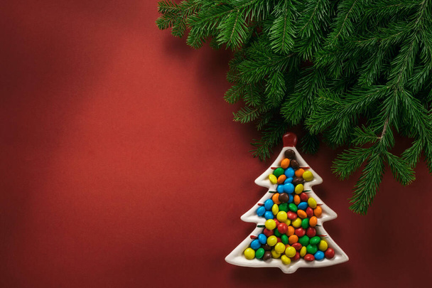 Christmas composition. A plate in the shape of a Christmas tree filled with colorful candies. Branches of spruce on a red background. Flat lay, top view, copy space. - Valokuva, kuva