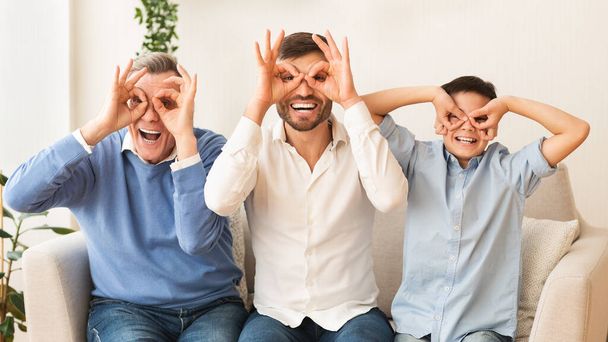 Grandfather, Father And Son Holding Fingers Near Eyes Sitting Indoor - Foto, immagini
