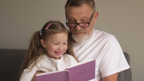 Grandfather teaches his granddaughter to read books. Family educational concept. An elderly man and a little girl leaf through a textbook - Кадри, відео