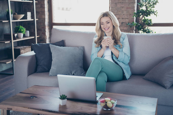 Portrait of her she nice attractive lovely charming cheerful cheery glad wavy-haired girl drinking coffee sitting on divan watching online video at industrial loft style interior room indoors - Foto, Imagen