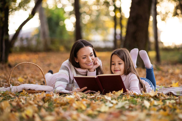 Μητέρα και κόρη απολαμβάνουν το φθινόπωρο στο πάρκο. Το κοριτσάκι μαθαίνει να διαβάζει.. - Φωτογραφία, εικόνα