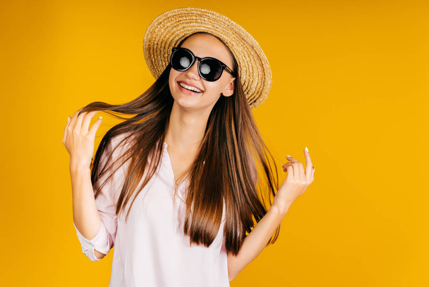 Girl in a hat and black glasses smiles joyfully and touches hair with her hands on a yellow background - Foto, immagini