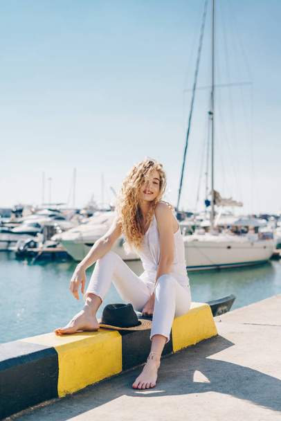 Lockige Blondine mit einem Lächeln in entspannter Pose sitzt auf der Brüstung der Seebrücke vor dem Hintergrund von Booten. Barfuß - Foto, Bild