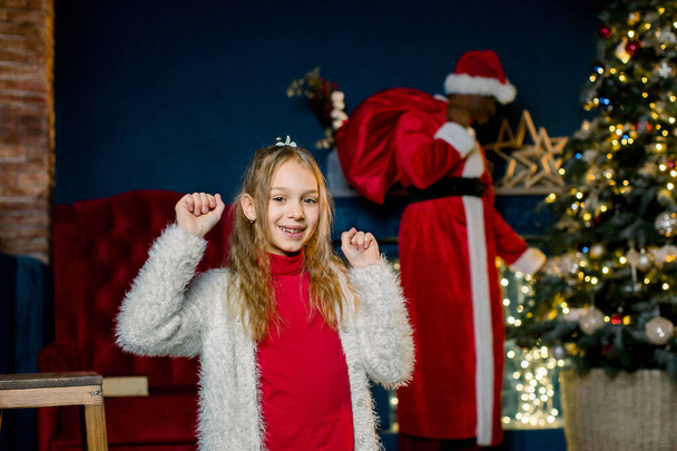 Little girl is happy that African Santa Claus came and puts presents at the Christmas tree - Valokuva, kuva