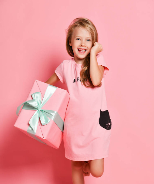 Christmas and New Year. Happy kid girl in pink dress with a big gifts box standing at pink wall holding hand at her ear - Фото, зображення