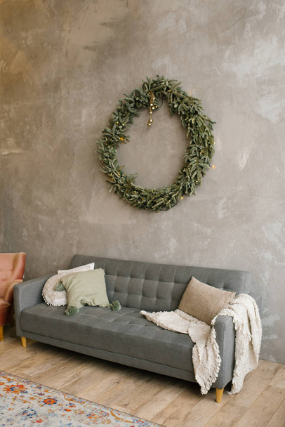 Gray sofa with pillows, over the sofa on the wall hangs a Christmas wreath. Scandinavian style in the living room - Photo, image