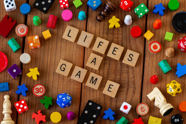 "Lifes a Game "spelled out in wooden letter tiles. Окруженный костями, картами и другими игровыми фигурами на деревянном фоне
 - Фото, изображение