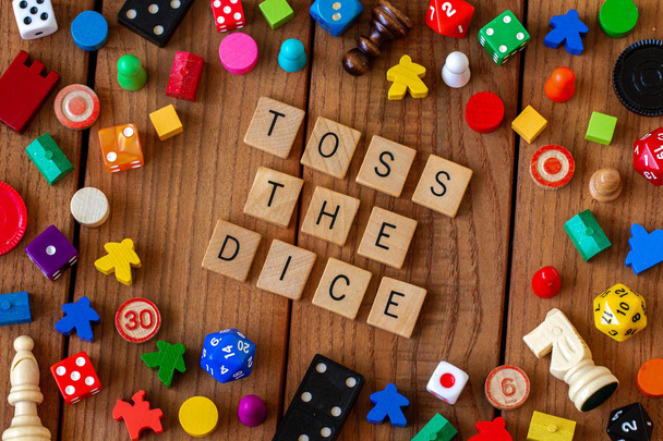 "Toss the Dice" spelled out in wooden letter tiles. Surrounded by dice, cards, and other game pieces on a wooden background - Photo, Image