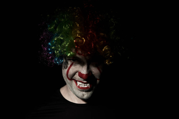 Nahaufnahme eines gruseligen Clowns, der aus der Dunkelheit hervorlugt. er trägt eine farbige Perücke und scharfe Zähne. - Foto, Bild