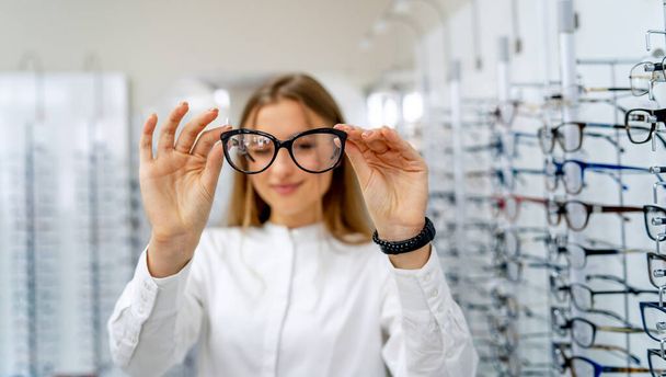female optometrist, optician is standing with many glasses in background in optical shop. Stand with spectacles. Eyesight correction. Woman holds spectacles in hands. Closeup. - 写真・画像